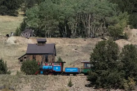 A small train passing by a rustic wooden building near a forest.