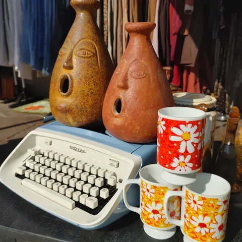 A vintage blue typewriter with face-shaped vessels and flower-patterned mugs on it.