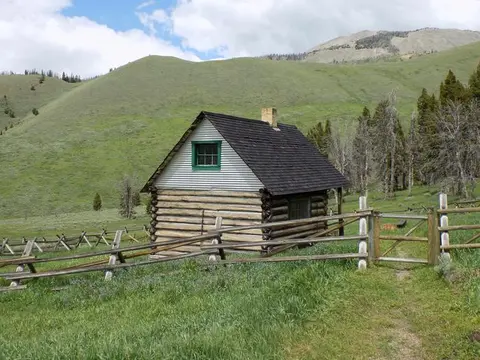 A small log cabin stands in a grassy valley with hills and trees in the background.