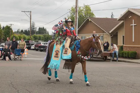 A woman in a parade outfit rides a decorated horse down a street.