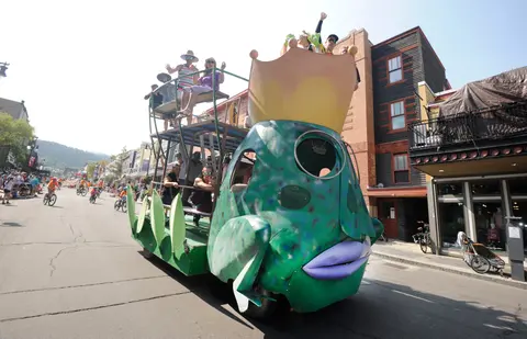 A decorated frog-shaped parade float travels down a street with people on board and crowds watching.
