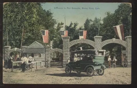 A vintage postcard showing the entrance to fair grounds with American flags and an early automobile.