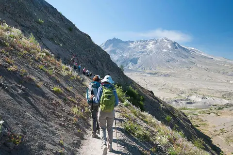 A group of hikers walking along a mountain trail with a large volcanic peak in the distance.