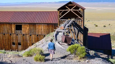 A person walks down a path near wooden historic buildings and a mining track structure in a landscape.