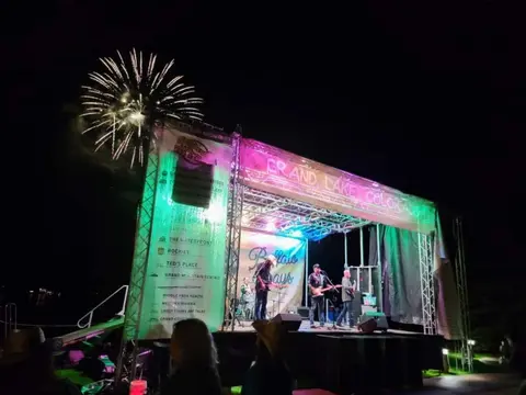 A band performs on a lit stage outdoors at night with fireworks in the sky.