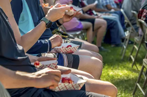 People sitting in folding chairs eating from small paper food trays outdoors.