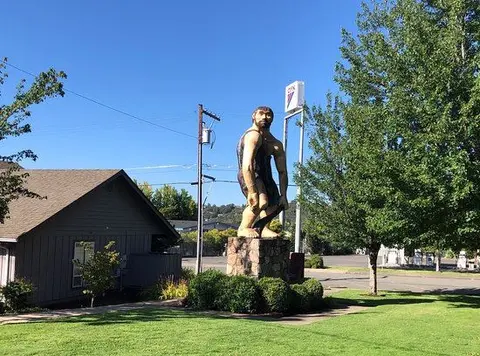 A large caveman statue stands on a stone pedestal outdoors near a building and trees.