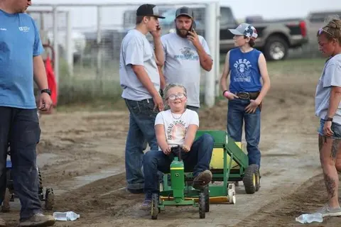 A child sits on a toy tractor while spectators watch outdoors.