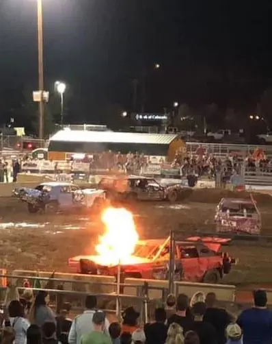 A car burns during a nighttime demolition derby event while spectators watch.