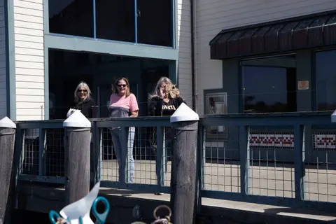 Three women stand on a pier by a building, with one taking a photo using a phone.