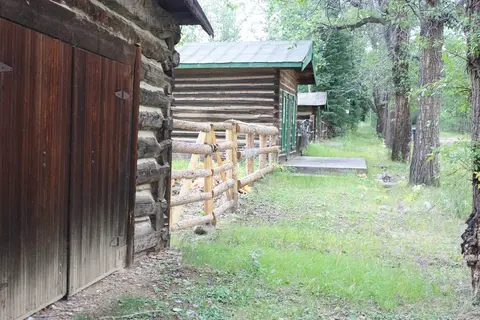 A row of rustic log cabins stands along a grassy path bordered by a wooden fence and trees.