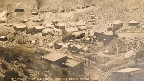 A historic black and white photo shows an elevated view of a settlement on a hillside.