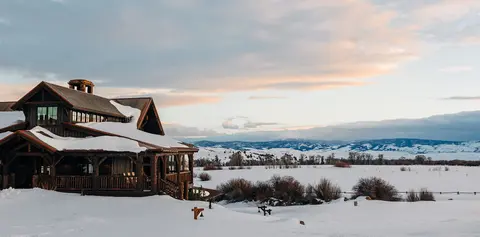 A large wooden lodge covered in snow with a snowy mountain landscape in the background.