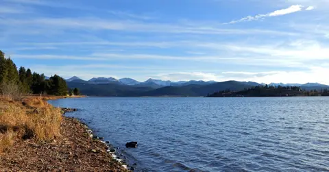 A scenic view of a large lake with distant snow-capped mountains and a treed shoreline.