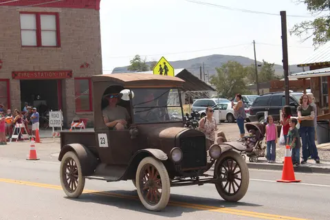 An old truck drives down a street during an outdoor event with people watching.
