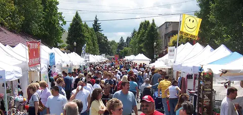 A crowded outdoor street fair featuring rows of white vendor tents under a bright sky.