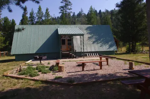 A green A-frame building sits in a wooded area with picnic benches outside.