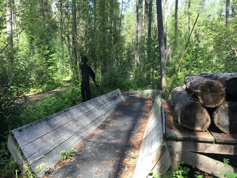 A paved path winds through a wooded area next to stacked logs and a silhouette statue.