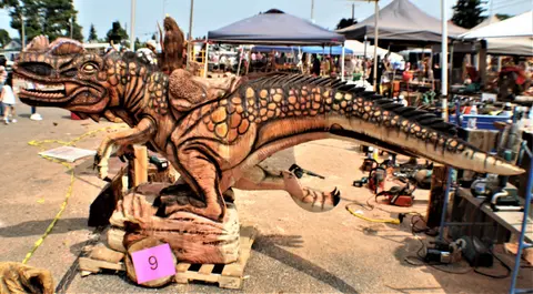 A wooden dinosaur sculpture displayed outdoors with canopy tents in the background.