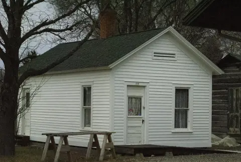 A small white wooden house with a dark shingle roof stands outdoors.