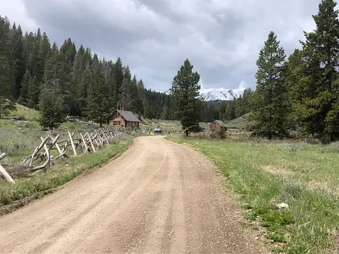 A dirt road leads through a valley with wooden cabins and pine trees under a cloudy sky.