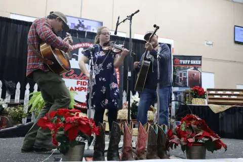 Three musicians play acoustic guitars and a fiddle on an indoor stage.