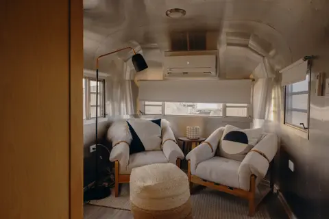 The interior of a retro-style silver trailer features two armchairs, a matching pouf, and modern lighting.