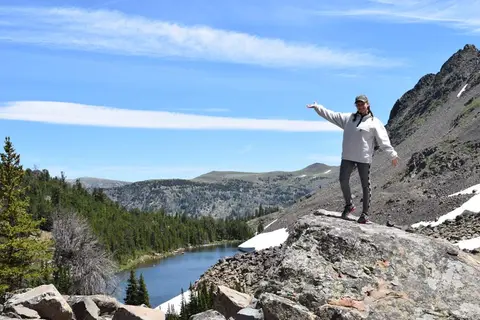 A hiker stands on a rocky outcrop with a mountain lake in the background.