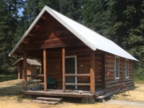 A rustic log cabin with a covered porch stands in a wooded area.