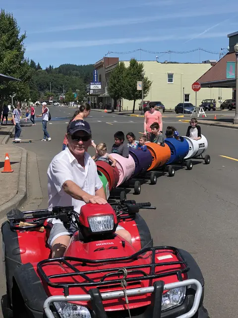 A man drives an ATV pulling a train of colorful barrel cars carrying children down a street.