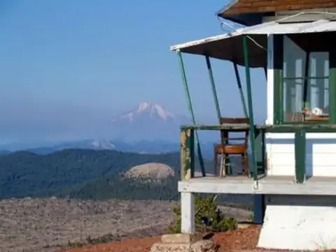 A lookout tower stands on a ridge overlooking a distant mountain and forested hills.