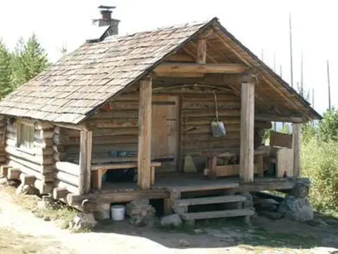 A rustic log cabin with a covered porch stands in a forest area.