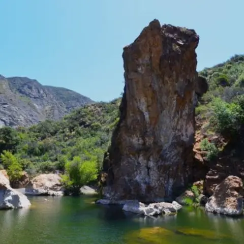 A tall rock formation stands beside a body of water in a mountainous landscape.