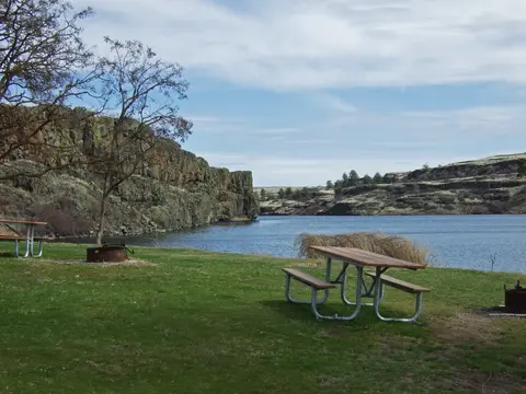 A scenic picnic area with tables by a lake and rocky cliffs.