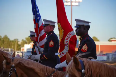 Marines in dress uniform carry flags while riding horses.