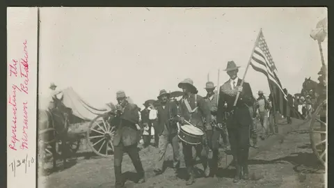 Historic black and white photograph of a group of people marching in a parade with an American flag and instruments.