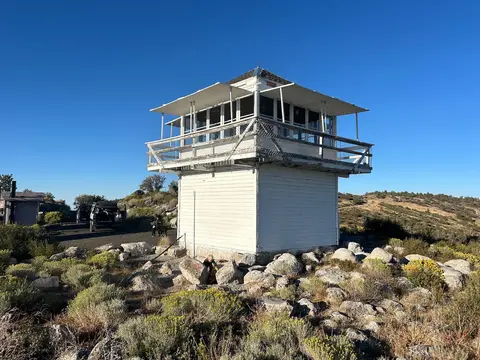 A white fire lookout tower sits atop a rocky hill under a clear blue sky.