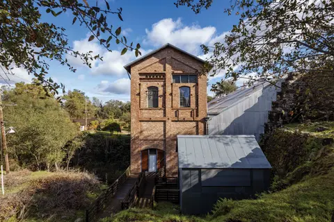 An old brick building with a metal roof stands outdoors near trees and foliage.