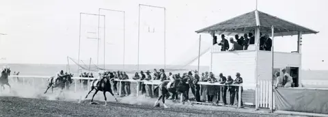 Black and white historic photo showing horses racing past a judges stand with spectators.