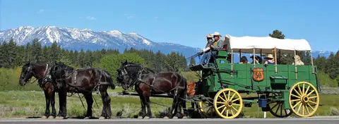 A team of four black horses pulls a green covered wagon with passengers in front of snow-capped mountains.