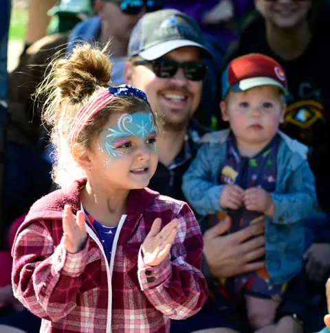 A young girl with face paint claps her hands in a crowd.