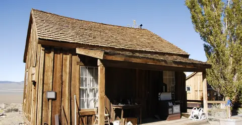 A rustic wooden cabin with a small porch sits in a dry landscape under a clear blue sky.