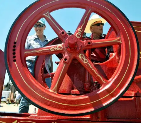 Two people stand next to a large red wheel on a machine.