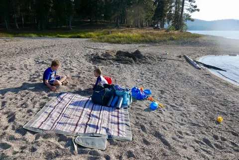 Two children play in the sand on a beach next to a lake.
