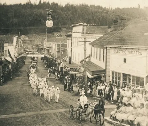 A historic photograph shows a parade proceeding down a street lined with buildings and crowds watching.