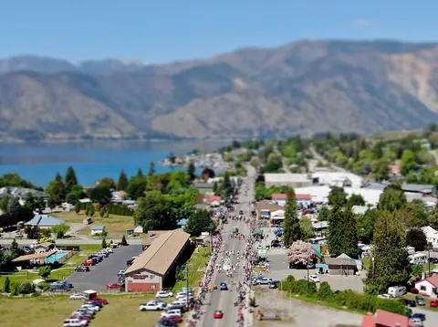 High-angle tilt-shift photograph of a town street with a crowd of people gathered along it, next to a lake and mountains.