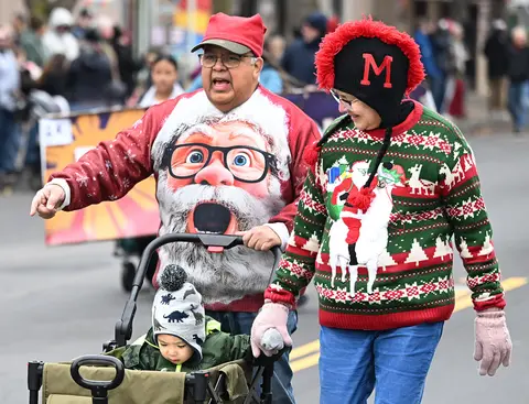 People in holiday sweaters and hats walk in an outdoor event with a child in a wagon.