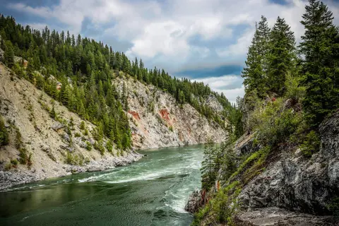 A wide river flows between steep, forested rocky banks under a cloudy sky.