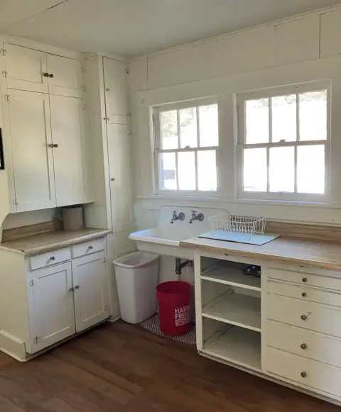 An interior shot of an old kitchen with white wooden cabinets and a sink.