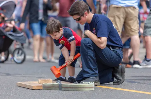 A young child hits a wooden block with a red mallet while a firefighter kneels nearby.